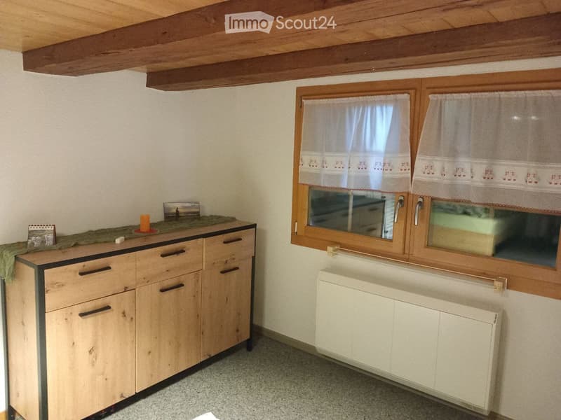 A small kitchen with wooden cabinets, a countertop, a radiator, and a window with curtains.
