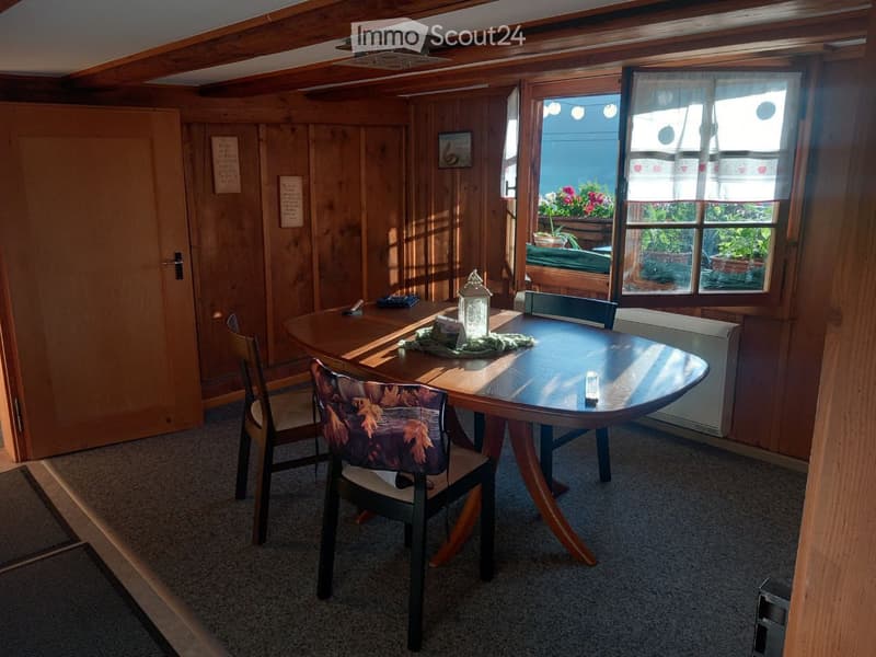 Cozy wooden-paneled room with a round wooden table, chairs, and a decorative lantern on the table. Large windows with white curtains let in natural light, and the room features exposed wooden beams on the ceiling.