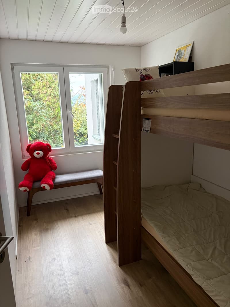 Cozy bedroom with a wooden bunk bed, a small bench with a red teddy bear, and a window with a view of greenery.