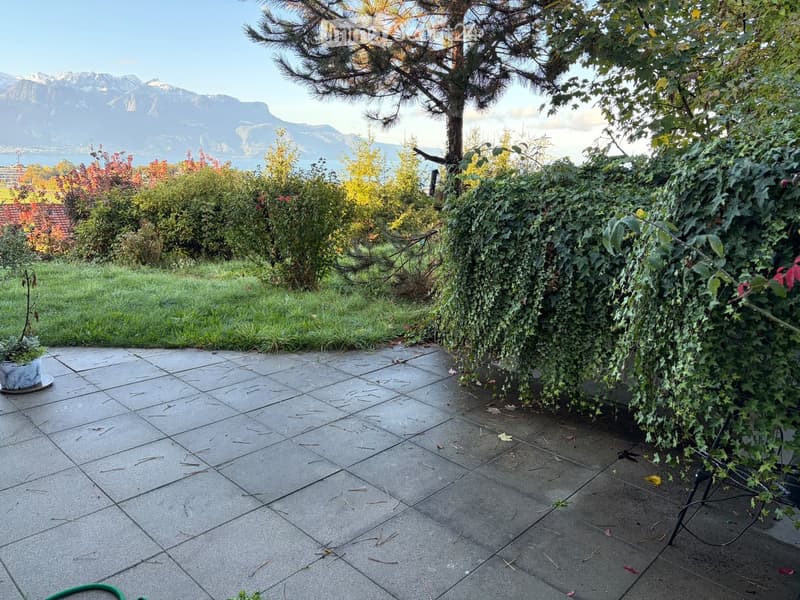 Garden area with a paved patio, surrounded by greenery and trees, with a view of mountains and a lake in the background.