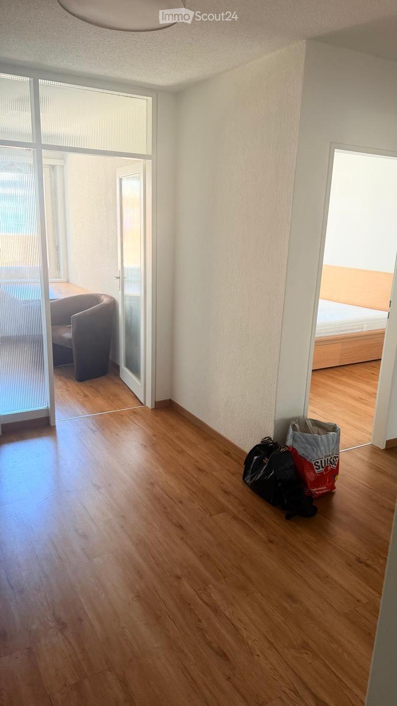 Empty room with wooden flooring, white walls, and large windows. Visible are a grey armchair, a bed, and some bags placed near the wall.