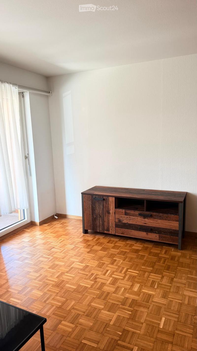 A modern, unfurnished room with wooden herringbone flooring, white walls, and a large window with white curtains. A dark wooden cabinet with drawers and a black table are visible.