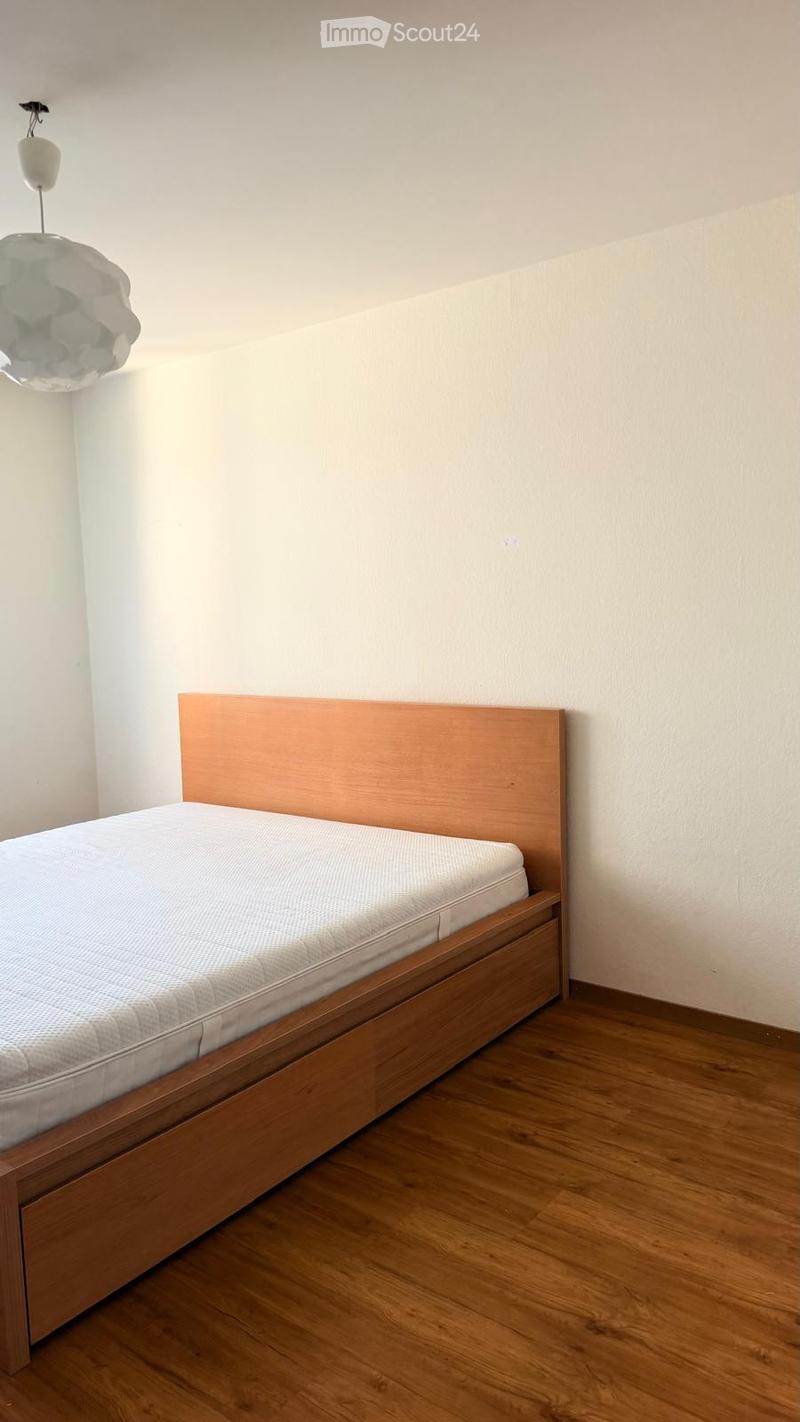 A simple bedroom with a wooden bed frame, a mattress, and a headboard. The room features light-colored walls and a wooden floor.