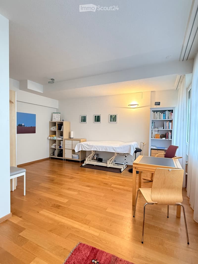 A well-lit room with wooden flooring, a massage table, a small table with chairs, shelves, and a cabinet.