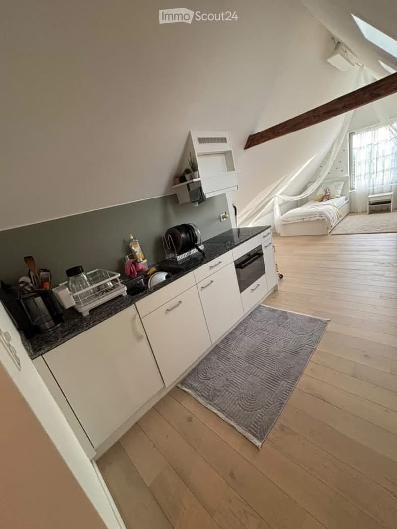 Open-plan attic space with a modern kitchen area and a bedroom area. The kitchen features white cabinets, a black countertop, and various appliances. The bedroom area includes a bed, a chair, and a patterned rug. Wooden floors and exposed beams are visibl