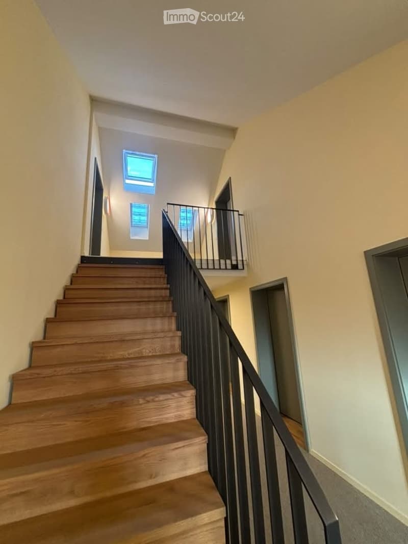 Wooden staircase with black metal railing leading to an upper level, featuring small windows and open doorways.