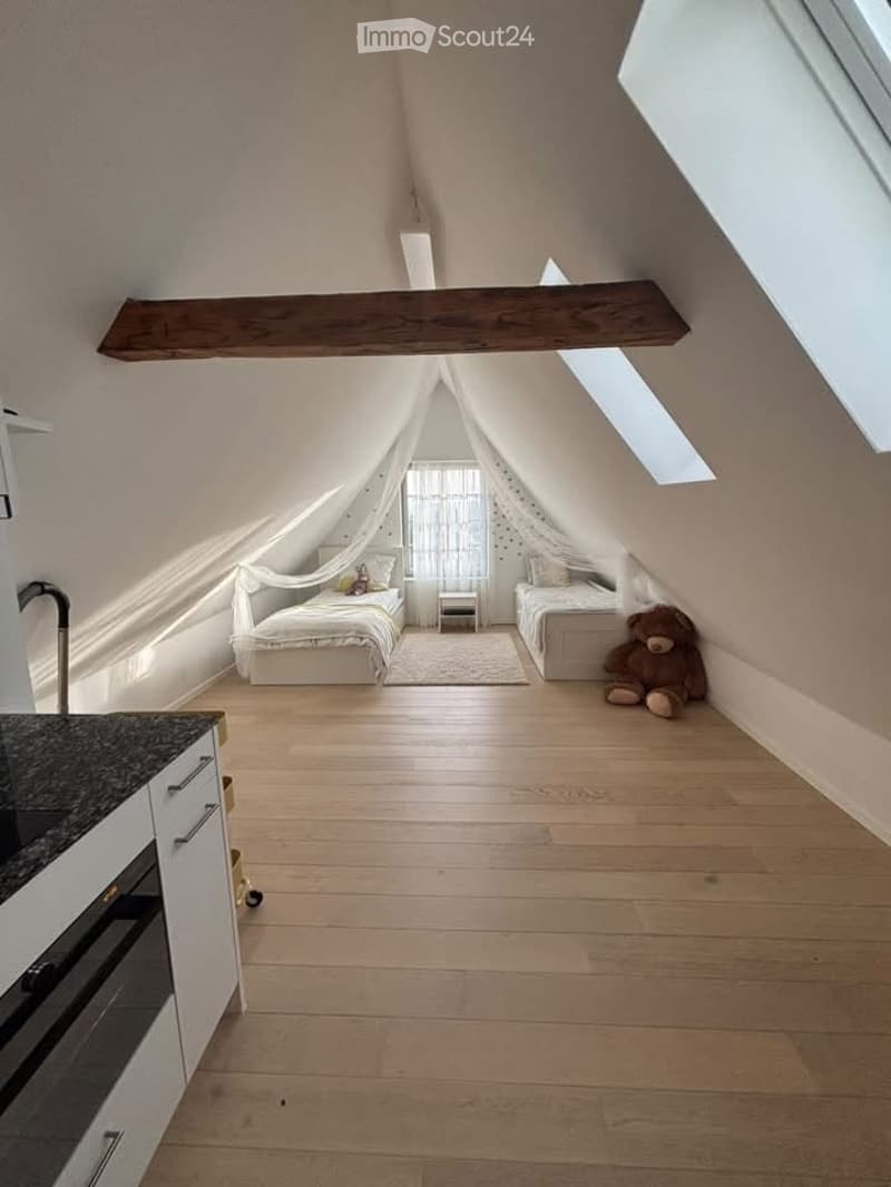 Loft-style bedroom with wooden floors, two single beds, nightstands, a small stool, and a kitchen area with an oven and faucet.