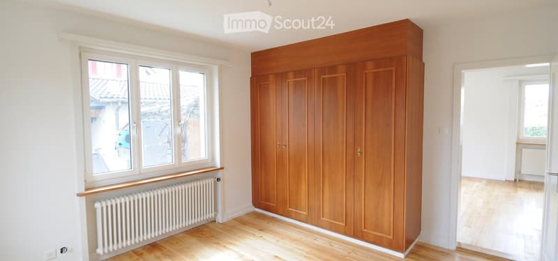 Empty room with wooden floor, large wooden wardrobe, white radiator, and large windows showing a view of a tiled roof and street.