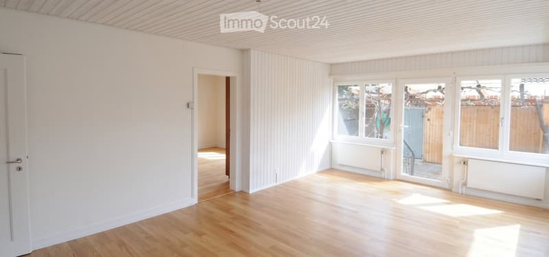 Empty room with wooden flooring, white walls, large windows, and a door leading to another space.