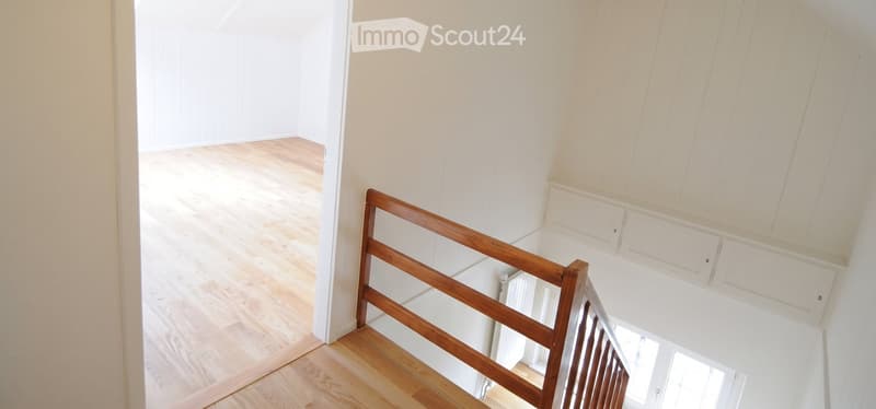 Empty room with light wooden flooring and white walls, featuring a wooden staircase railing and a visible doorway leading to another space.