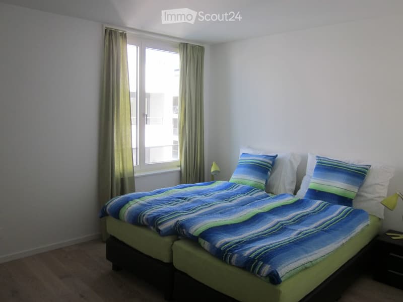 Bedroom with a double bed, blue and green striped duvet, white pillows, nightstand with lamp, and window with curtains.