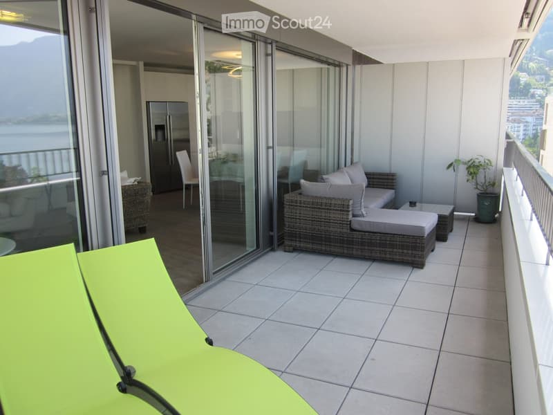 Balcony with tiled floor, modern furniture including a sofa, chairs, and a table, with a scenic view of mountains and water through large glass doors.