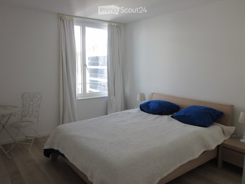 Bedroom with a wooden bed, blue pillows, nightstand, small table, and chair near the window with curtains.