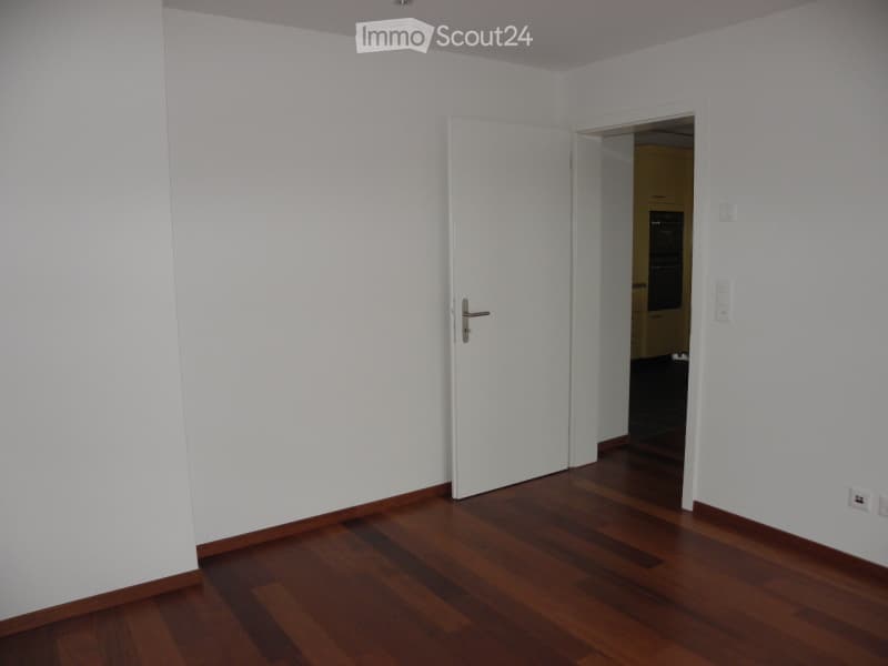 Empty room with white walls and wooden flooring, featuring an open door leading to another room.