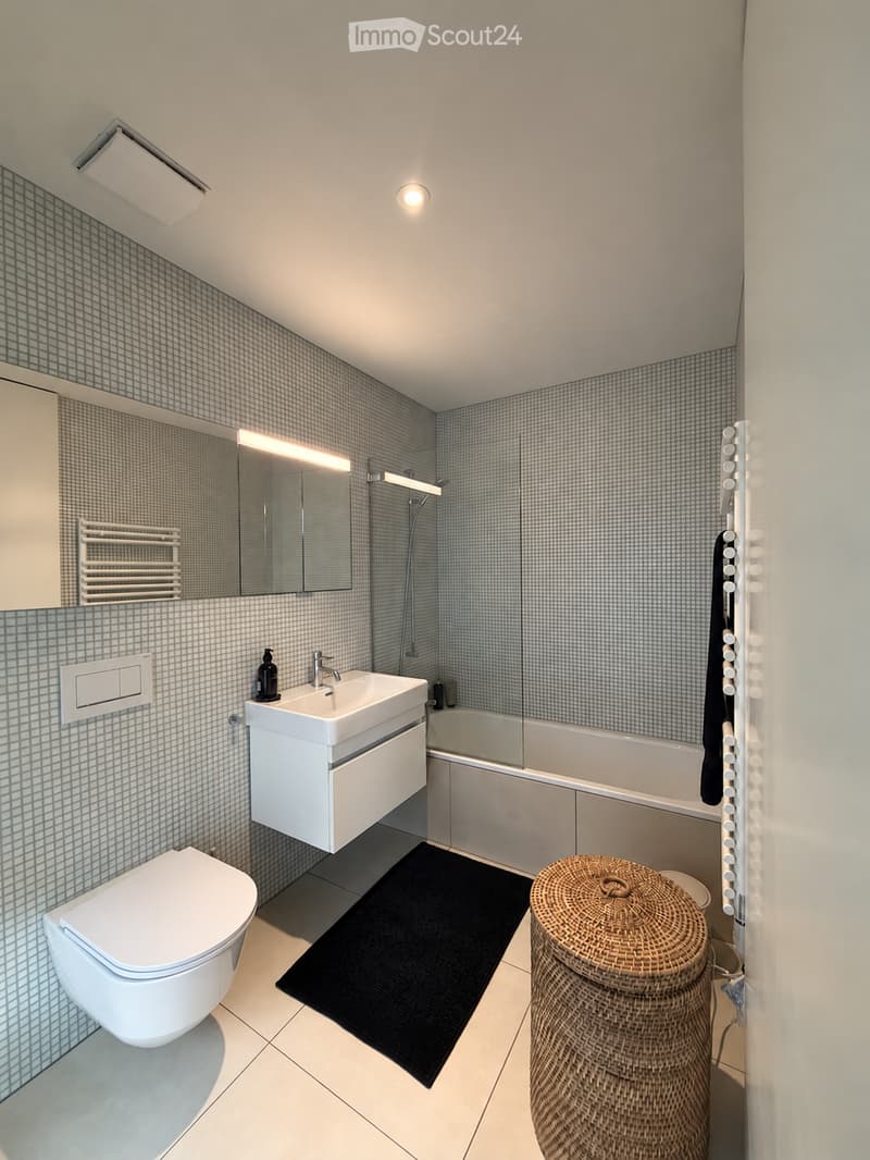 Modern bathroom with white tiles, a sink with a mirror, a toilet, a bathtub, a wicker basket, and a radiator.