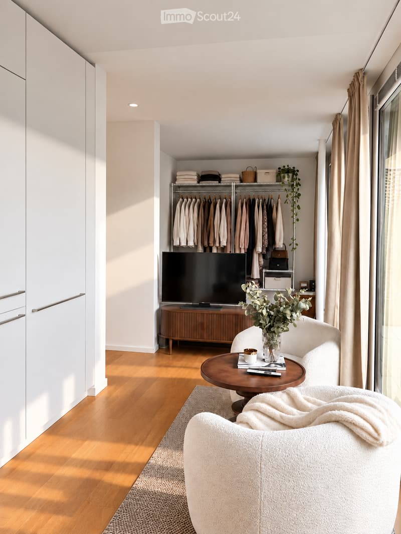 Modern living room with a white sofa, wooden coffee table, flat-screen TV, and a wardrobe with hanging clothes.