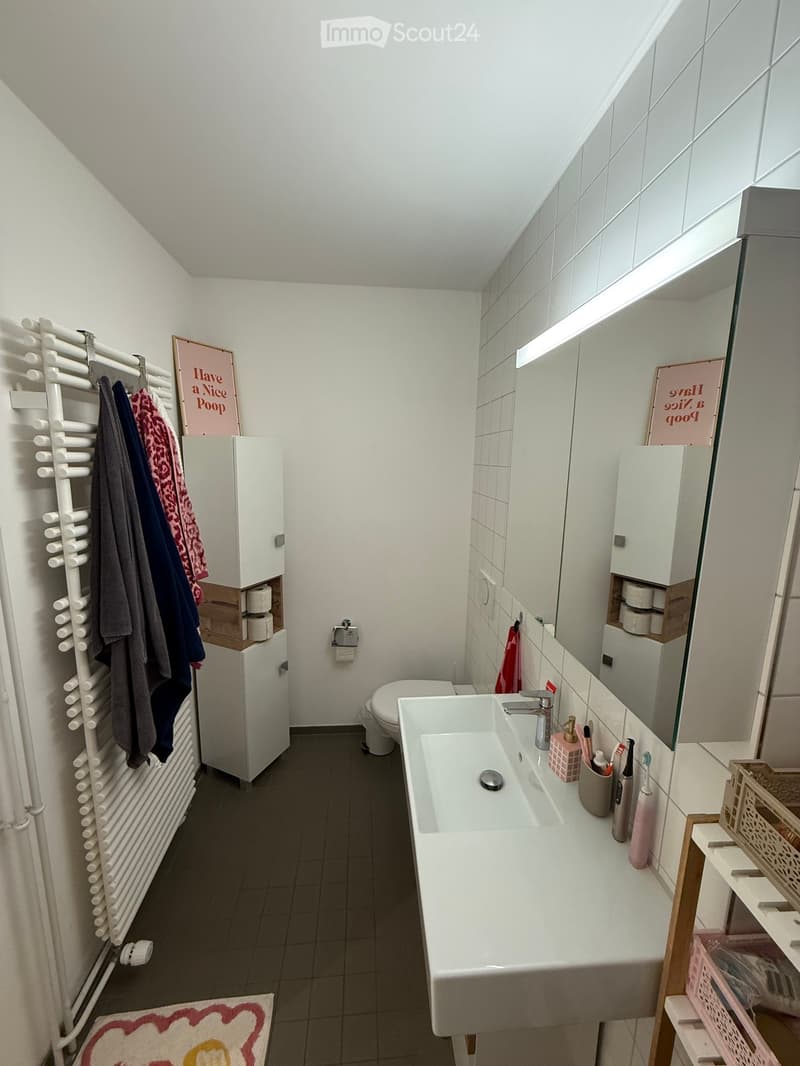 Modern bathroom with white tiles, a large sink with a mirror, a toilet, a towel rack with towels, and a cabinet with toilet paper. The sink has a faucet and various toiletries on the counter.