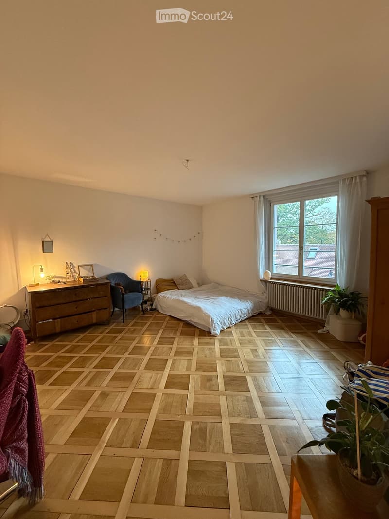 Cozy bedroom with wooden herringbone flooring, a large bed with white bedding, a blue armchair, a wooden dresser with a lamp, and a window with sheer curtains.