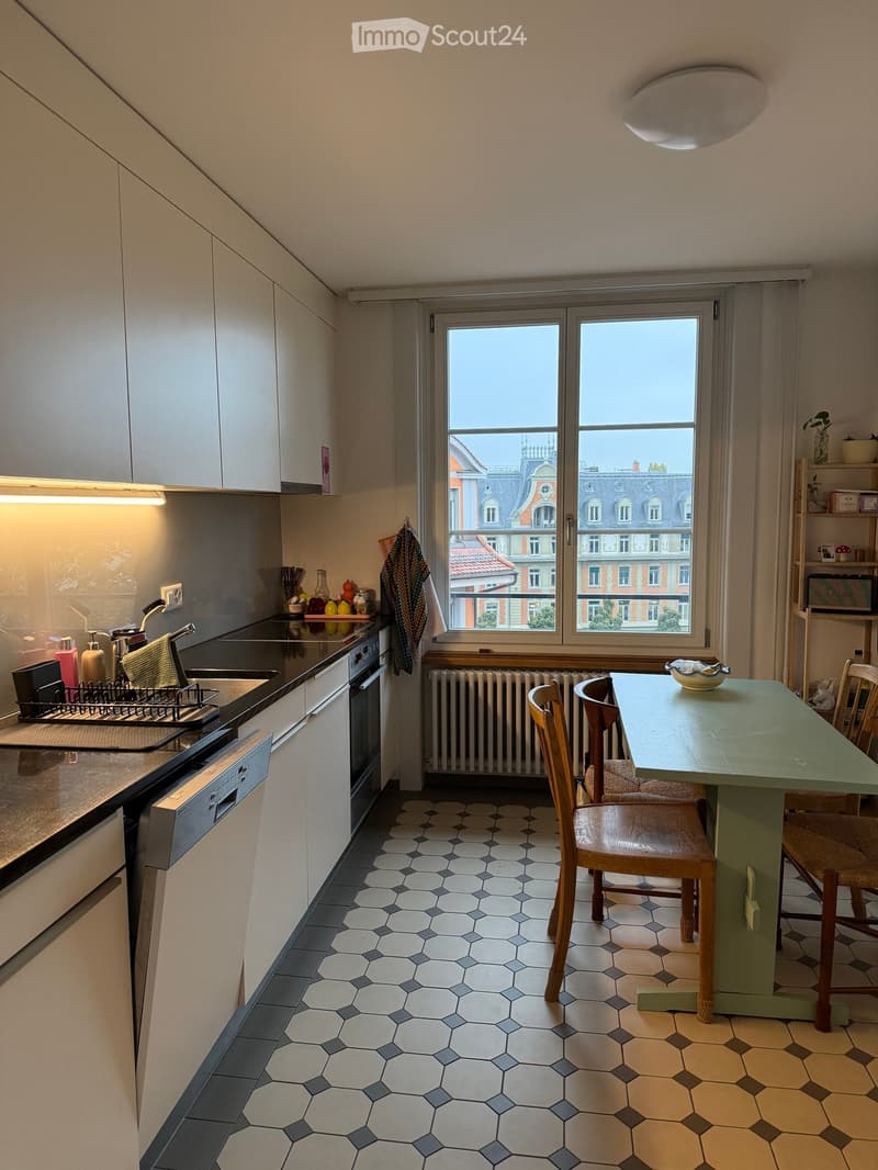 Modern kitchen with white cabinets, stainless steel appliances, and a green dining table with wooden chairs. The room features a large window with a city view, a radiator, and a patterned tile floor.