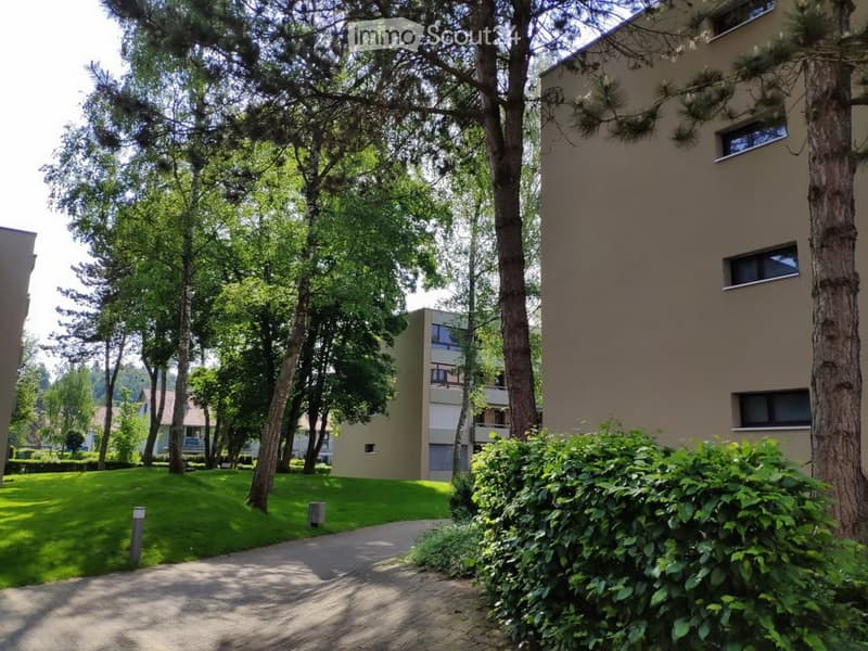 Modern multi-story residential building surrounded by lush greenery and trees, with a well-maintained lawn and pathway leading to the entrance.