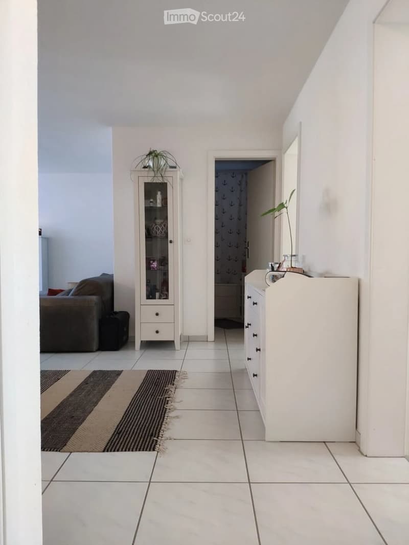 A modern hallway with white tiled flooring, a beige and brown rug, a white cabinet with glass doors, a small plant on top, and a sofa visible in the adjacent room.