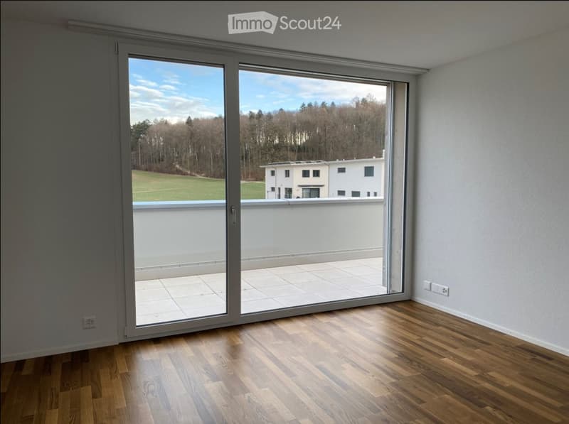 Empty room with wooden flooring, large sliding glass doors leading to a balcony, and a scenic view of a green field and trees.