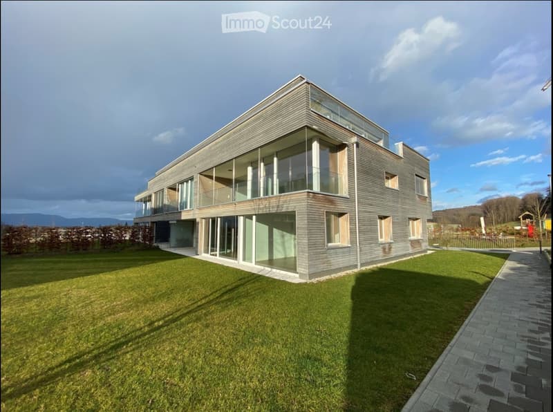 Modern two-story house with grey wooden facade, large windows, and a raised ground floor. Features a spacious lawn and a paved pathway leading to the entrance.