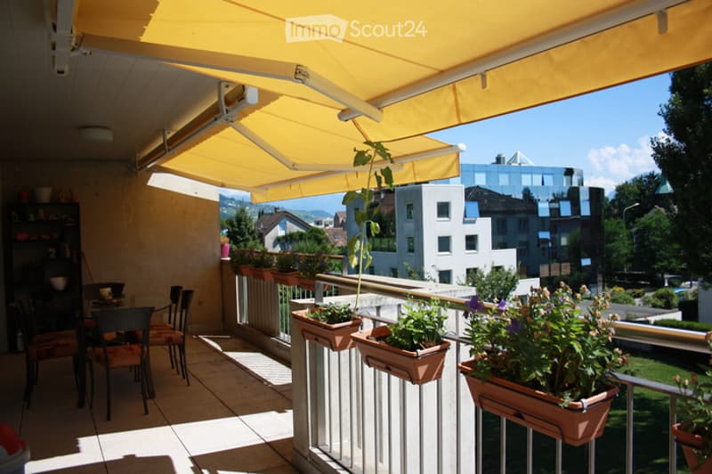 Balcony with a yellow retractable awning, several potted plants, and a small dining set with chairs and a table.