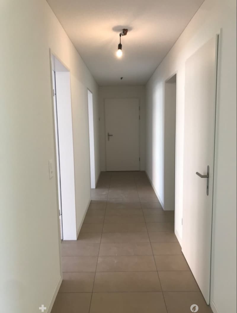 Empty hallway with beige tiled floor, white walls, and a single ceiling light fixture. Doors are visible on both sides.