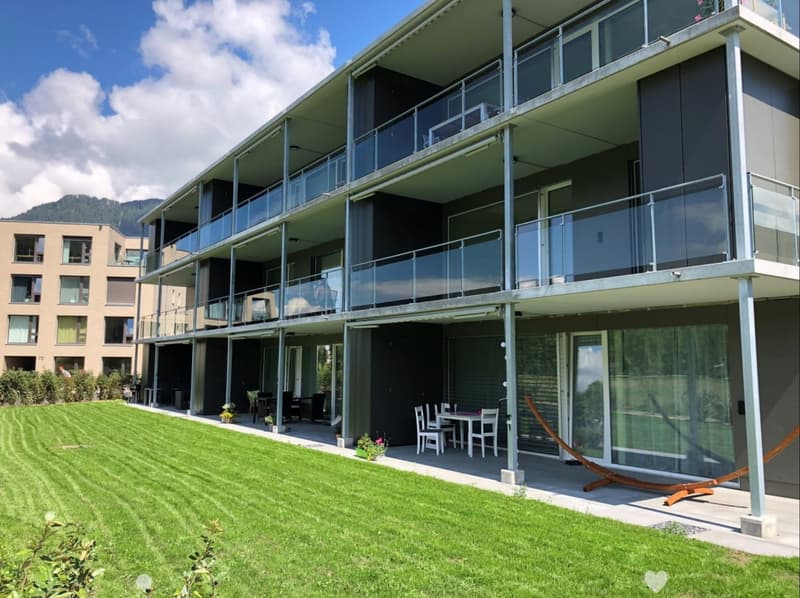 Modern multi-story building with balconies and a well-maintained lawn in front. Outdoor seating area with a table, chairs, and a sun lounger.