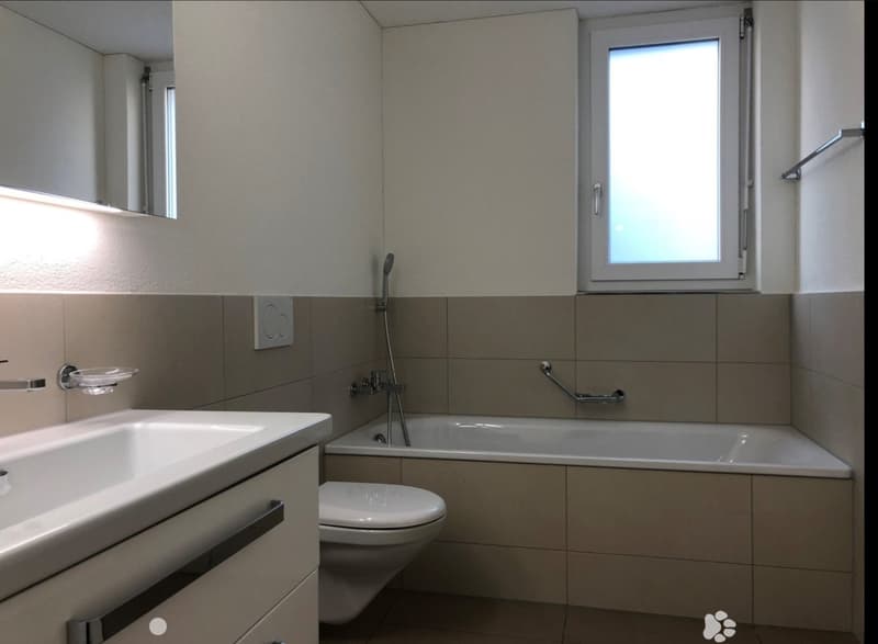Modern bathroom with a white sink, toilet, bathtub, and a large mirror above the sink. Features a window with frosted glass and a handheld showerhead.