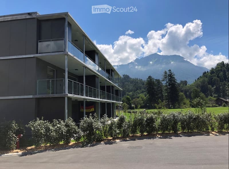 Modern two-story building with balconies, surrounded by greenery and mountains in the background.