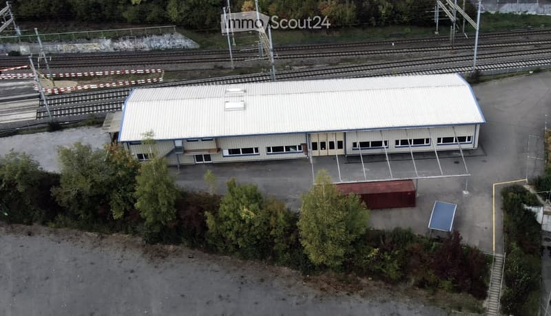 A single-story industrial building with a metal roof and multiple windows, situated near railway tracks. The surrounding area includes trees, a small container, and a blue structure.