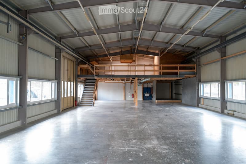 Large, empty industrial-style space with concrete floors, metal walls, and a wooden staircase leading to an upper level. Natural light streams in from large windows on the left side.