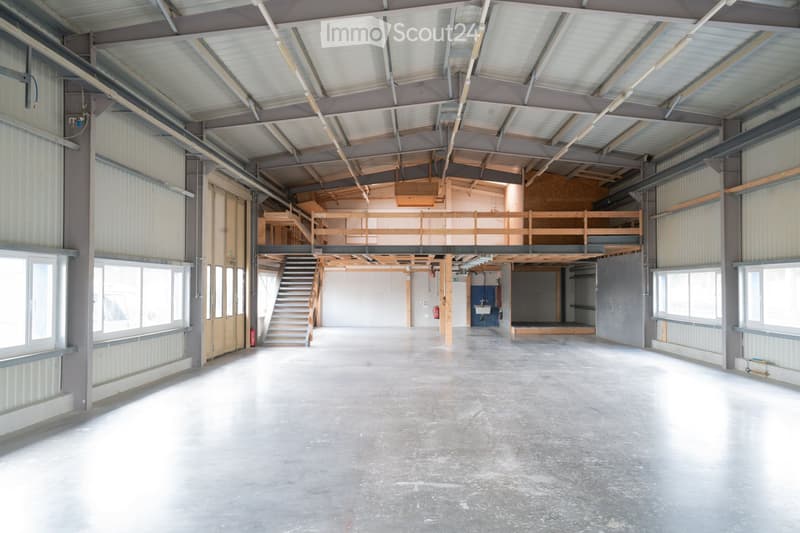 Large, open industrial-style space with concrete floors, metal walls, and exposed wooden beams. Features include a staircase leading to an upper level, large windows, and a small wash area with a sink.