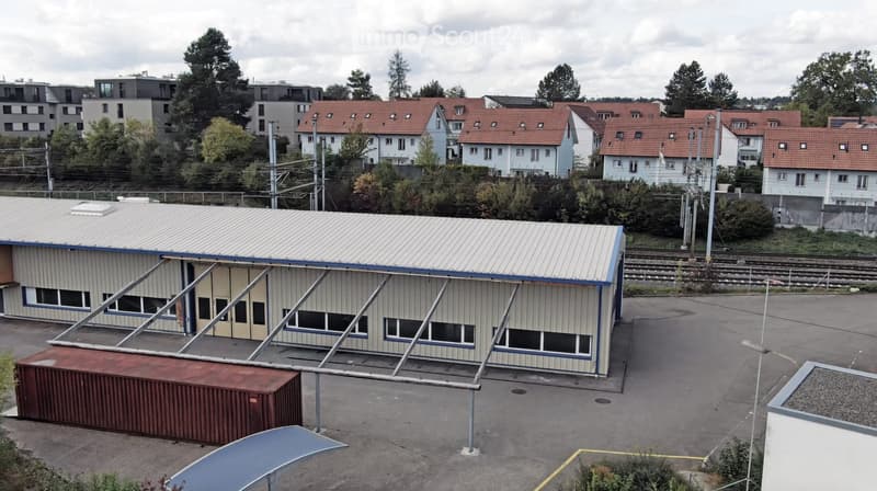A modern industrial-style building with a metal roof and large windows, situated in an urban area with surrounding residential houses and trees.
