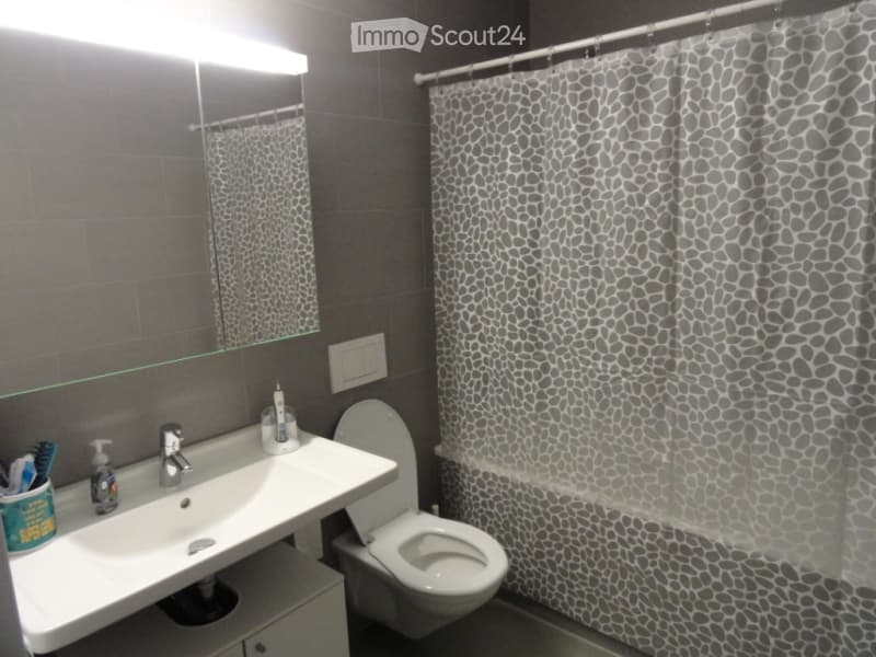 Modern bathroom with a white sink, chrome faucet, mirror, toilet, and a shower curtain with a grey and black pattern.