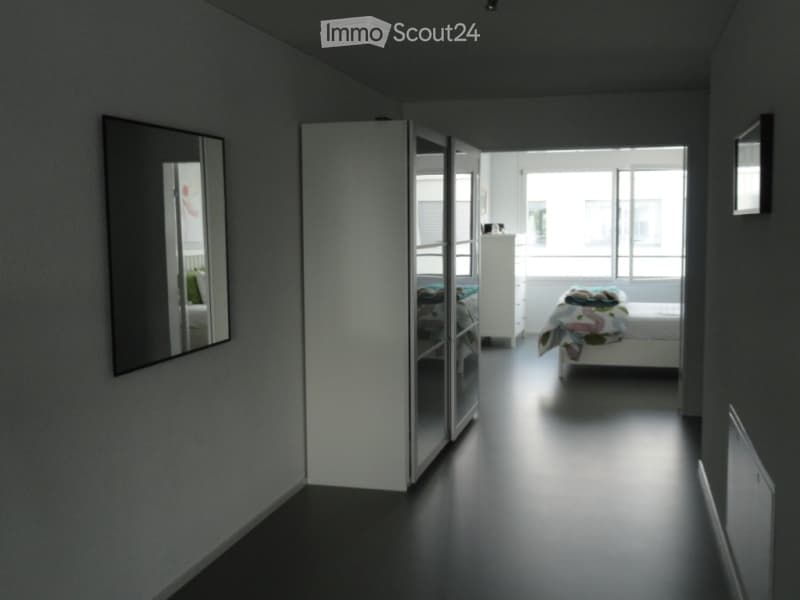 Modern, minimalist bedroom with dark flooring, large mirror, sliding wardrobe, and neatly made bed.