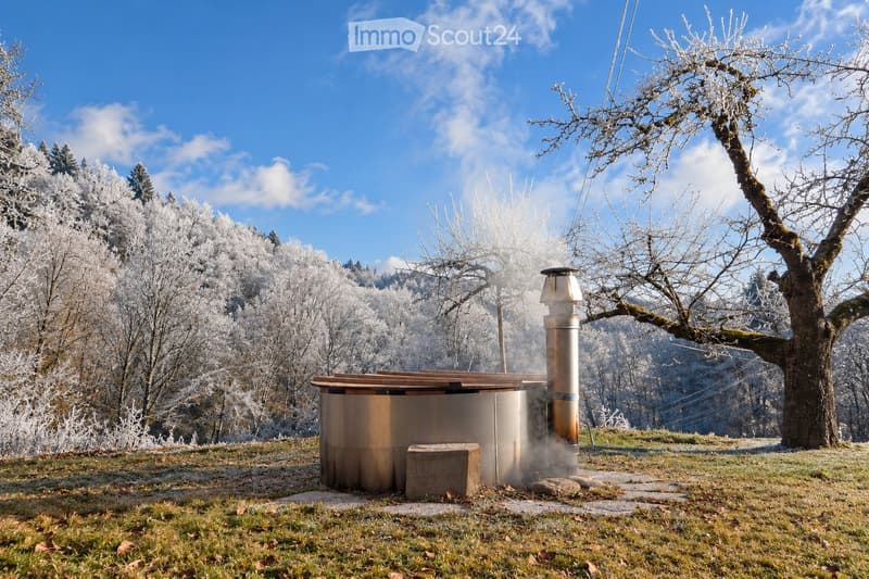 A wooden hot tub with a metal chimney is situated in a grassy area surrounded by frost-covered trees and a clear blue sky.