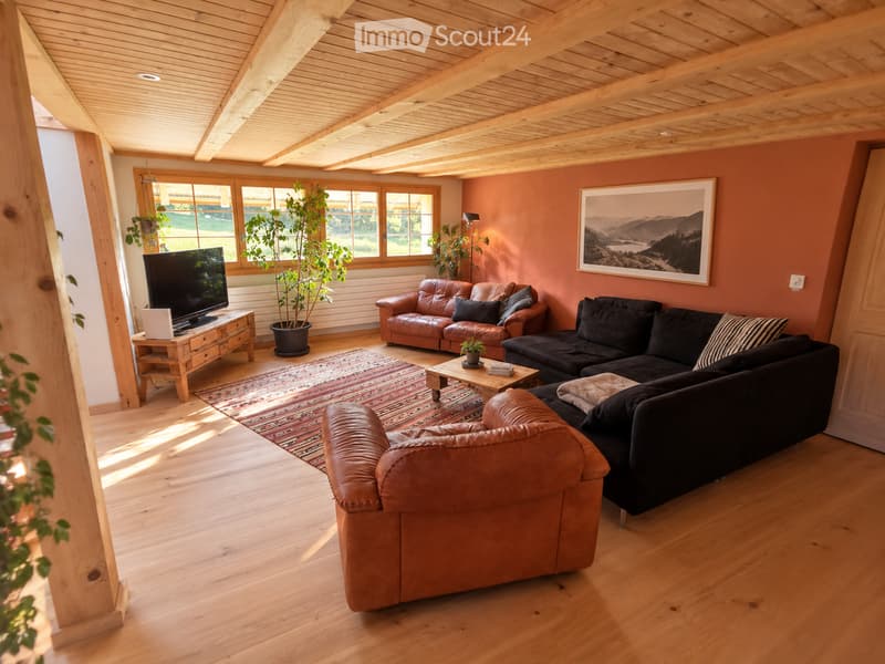 Bright living room with wooden beams, hardwood floors, and large windows. Features a black sectional sofa, brown leather armchair, coffee table, TV on a wooden stand, and a potted plant.