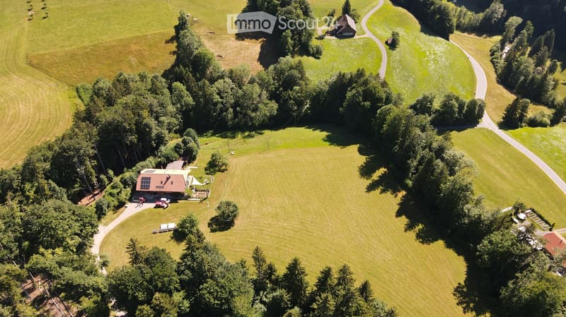 Aerial view of a rural property with a house, parking area, and surrounding green fields and trees.