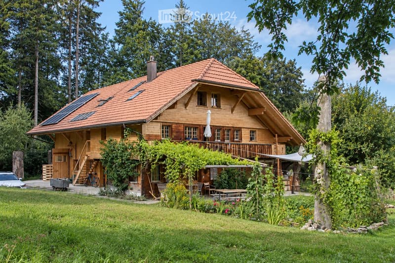 A two-story wooden house with a red-tiled roof, surrounded by a lush garden and trees. Features include a balcony, solar panels on the roof, and a small patio area with outdoor furniture.