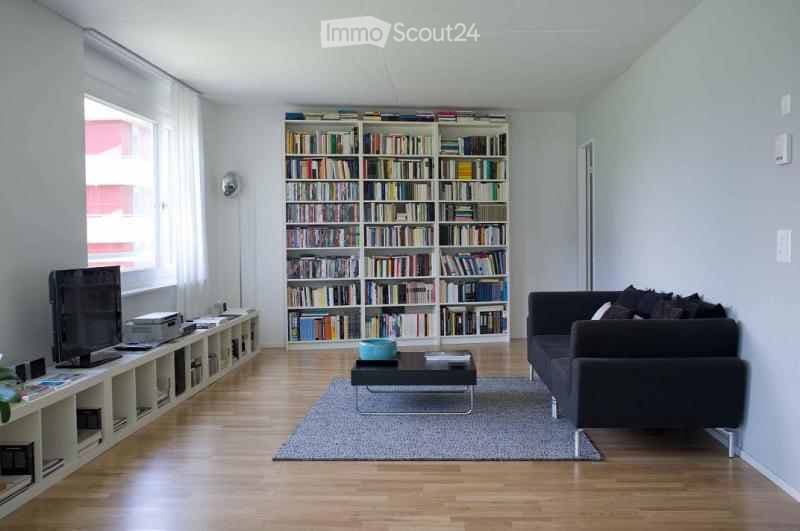 Modern living room with a large bookshelf filled with books, a black sofa, a coffee table, a television on a stand, and a floor lamp near the window.