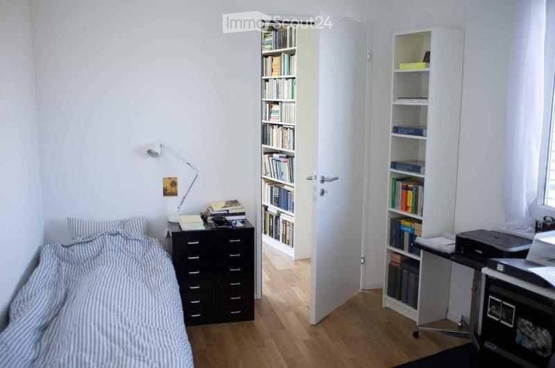 Cozy bedroom with a bed, nightstand, lamp, bookshelf, desk, and chair. The room features light wooden flooring and white walls.