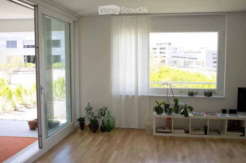 Bright, modern living room with large windows and a view, featuring a white shelf unit with books and decorative items, several potted plants, and glass vases near the window.