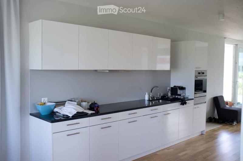 Modern kitchen with white cabinets, black countertop, and stainless steel appliances. Features a sink, faucet, oven, and a small stool near the window.
