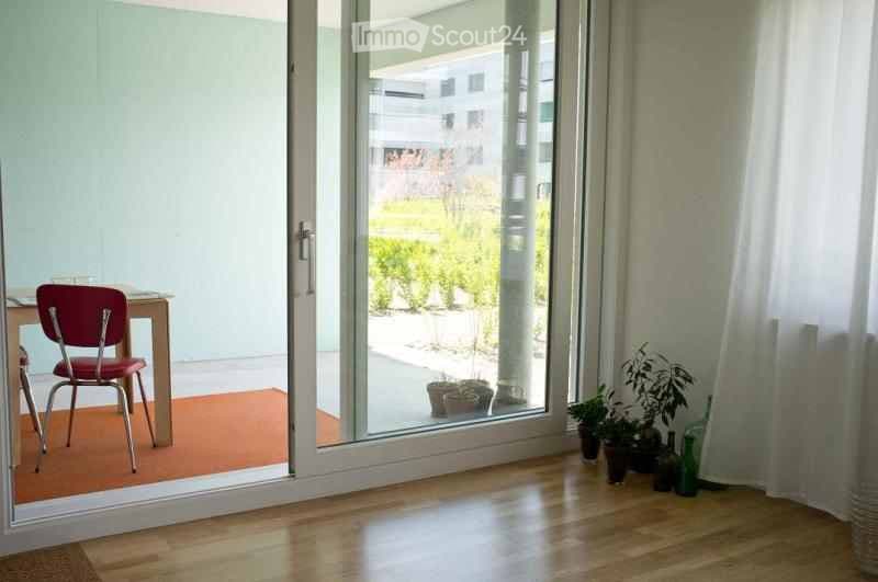 A small, well-lit room with wooden flooring, a red chair, a small table, and several potted plants near the sliding glass door that opens to a garden area.