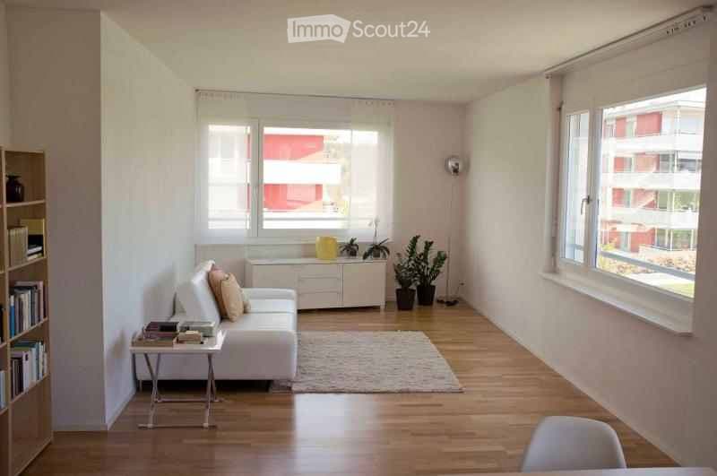 Bright, modern living room with white walls, wooden flooring, large windows, a white sofa with cushions, a coffee table, a bookshelf, and potted plants.