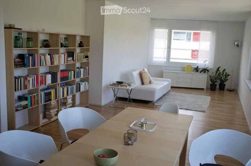 Bright, open living and dining area with white walls, wooden flooring, and large windows. Features a white sofa with pillows, a small coffee table, a bookshelf filled with books and decorative items, a dining table with chairs, and a bowl on the table.
