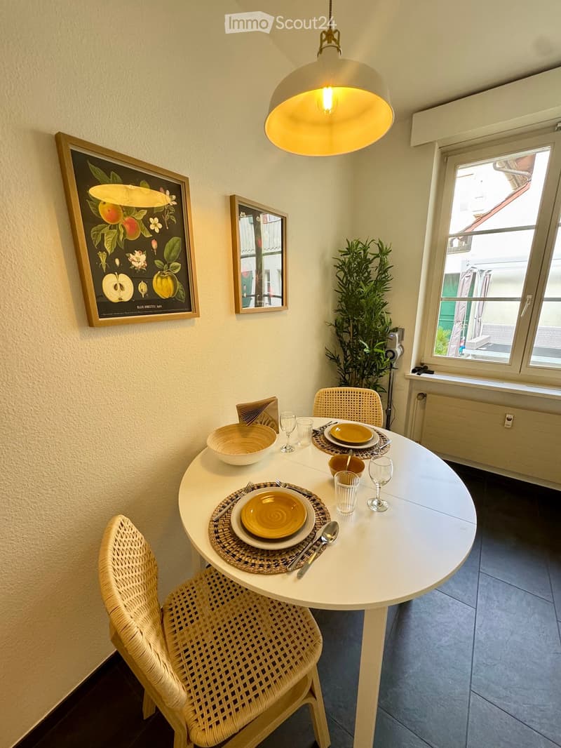 A small, cozy dining area with a round white table set for four, featuring woven placemats, yellow plates, bowls, and wine glasses. Two wicker chairs are visible, along with a framed picture and a potted plant near the window.
